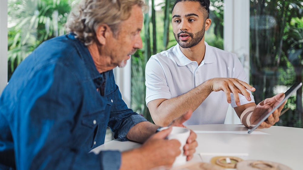 An olderly man with coffee and a young man with a tablet sit at a table and talk