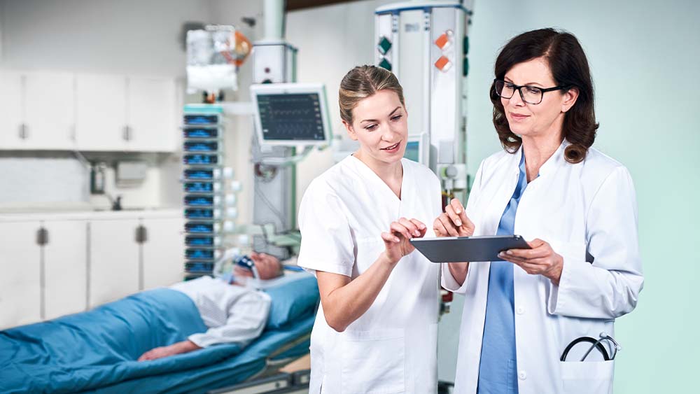 A doctor and a nurse are discussing the dosage for B. Braun smart infusion, which is being directly input from a tablet. The patient in the ICU unit can be seen in the background, connected to a tower full of smart infusion devices.