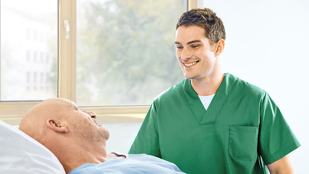 Male nurse smiling at a patient lying in the bed representing “More time for patient care”. 